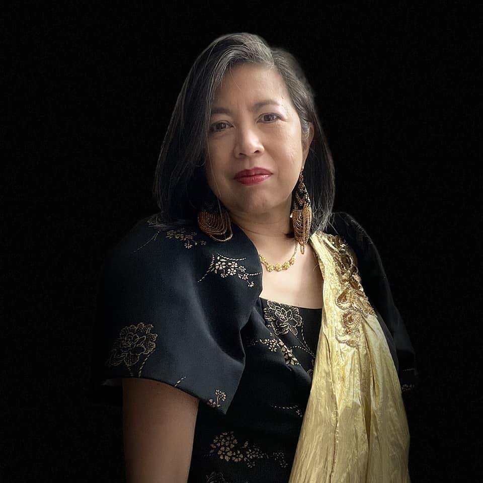 Emily P. Lawsin, Filipina woman with shoulder-length black hair with gray highlights, wearing a black butterfly sleeve gown with gold flower design and gold sari, gold necklace, and black and gold dangling beaded earrings, in front of a black background.